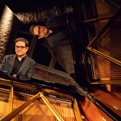 Pianist Coady Green in dark blazer and glasses poses seated at an open grand piano, under warm stage lighting.