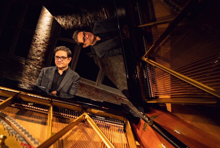 Pianist Coady Green in dark blazer and glasses poses seated at an open grand piano, under warm stage lighting.