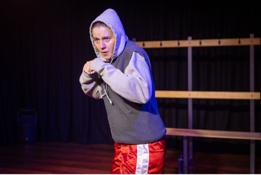 A scene from the play Bitch Boxer, featuring a performer in red boxing shorts and a hoodie shadow boxing on the stage.