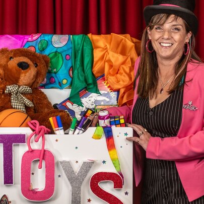 A woman in a pink blazer posing next to an overflowing toy box in front of a red stage curtain.