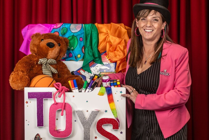 A woman in a pink blazer posing next to an overflowing toy box in front of a red stage curtain.