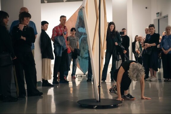 Dancer bending beside an artwork in a gallery surrounded by onlookers.