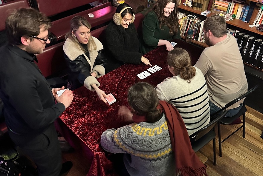 Looking down at a table of 6 people performing card tricks with another man looking on. 