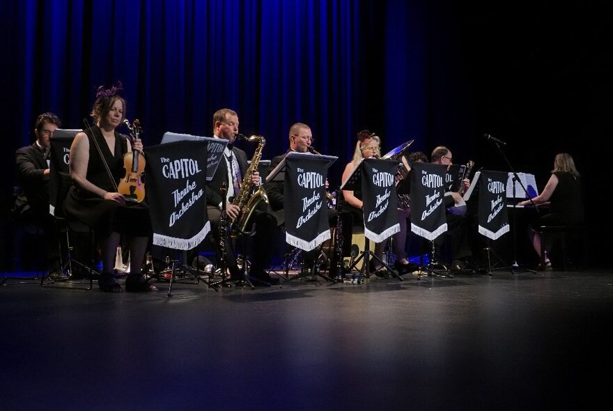 Mixed band of seated musicians performing on stage with banners.