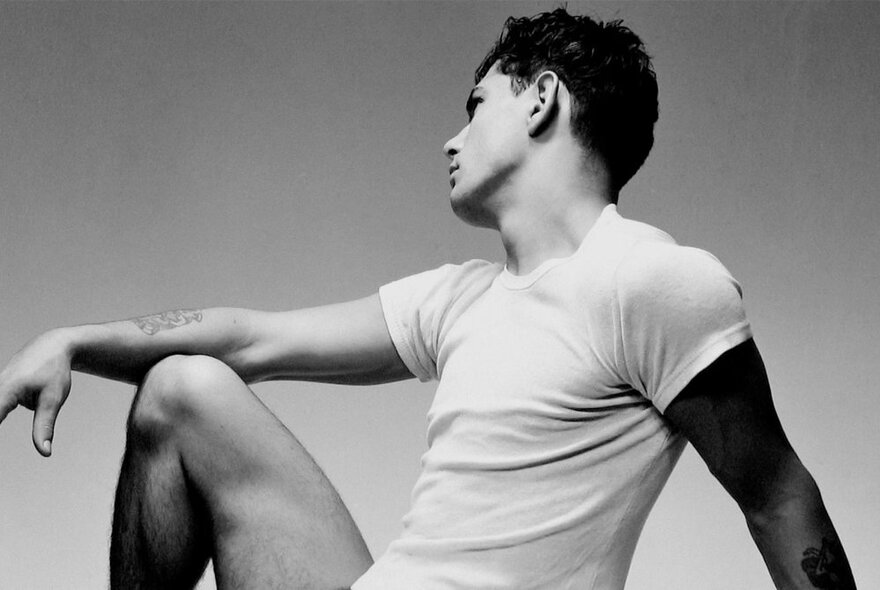 A black and white image of a man in a white t-shirt posing elegantly, his head turned away and an arm resting on his knee.