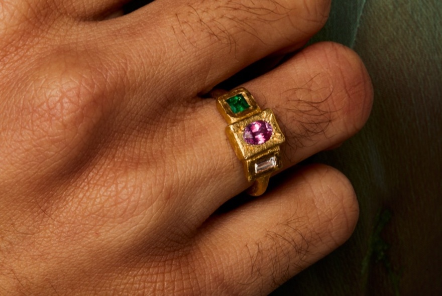 A hand with hairy fingers and a ring with several coloured block stones. 