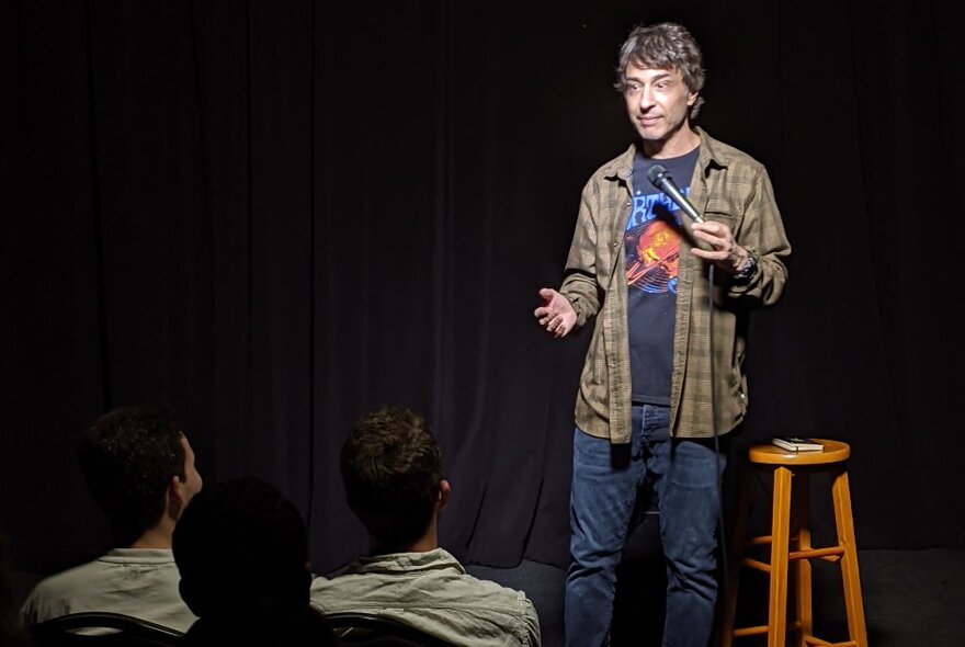 Comedian Arj Barker, on stage with a wooden stool next to him, audience seen from behind.