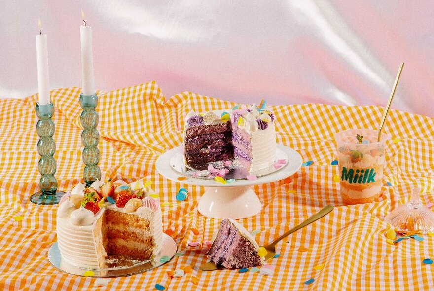 A surface with a yellow gingham tablecloth with layer cakes on stands, two candlesticks in green holders and a clear drink cup with a stray