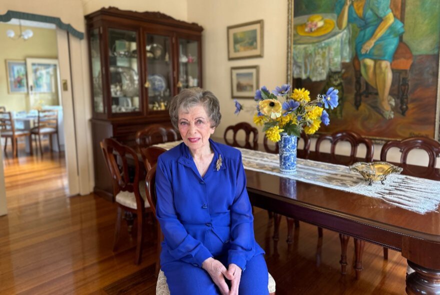 An elderly woman with short gray hair, wearing a vibrant royal blue long-sleeved dress, sitting on a chair in a dining room, with a glass fronted display cabinet in the background, and paintings on the wall.
