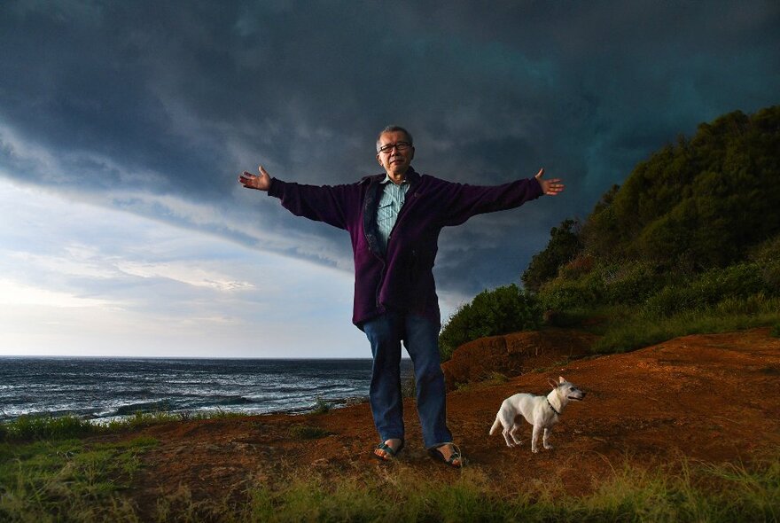 William Yang, a man standing in a coastal landscape with his arms stretched out by his side, a small white dog next to him.