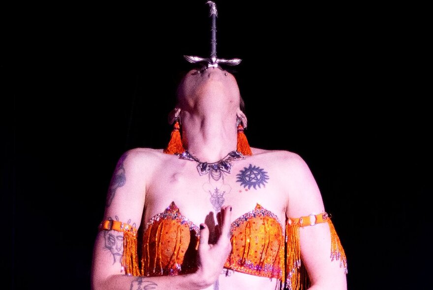 A tattooed woman in an orange sequined bikini top leaning her head right back with the handle of a sword showing above her mouth.