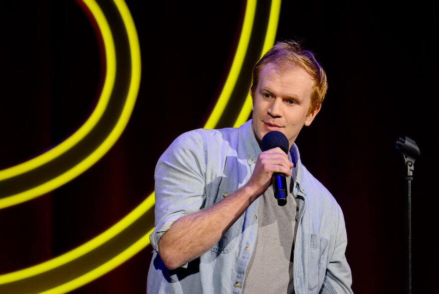 A comedian performing stand-up on stage holding a microphone in front of a black backdrop with yellow curved lines.