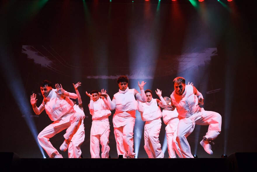 A large group of dancers from the Royal Family Dance Crew, in white tops and white pants perform a dance routine on a dark stage with dramatic spotlight lighting.