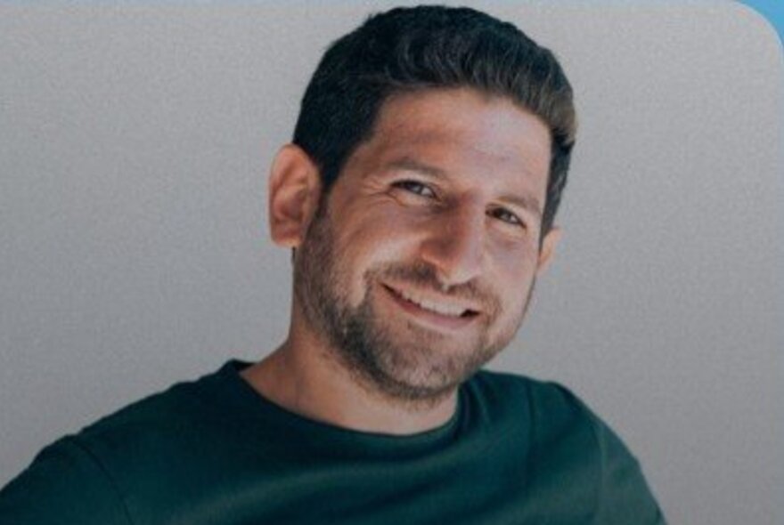 Portrait shot of Syrian comedian, Bahaa Dabbagh, wearing a green t-shirt and smiling at the camera; grey background.