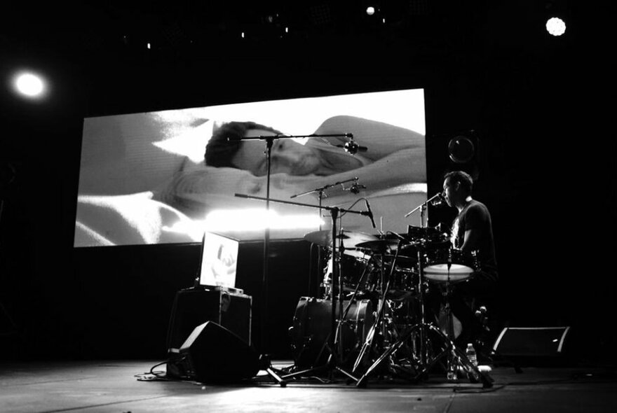 A solo jazz drummer on a darkened stage with a projector screen behind him showing a scene from the movie, Birdman.