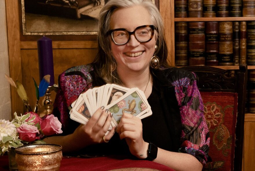 Smiling person wearing glasses holding up a deck of fanned out tarot cards in her hand, behind her is a bookshelf with leather bound books.