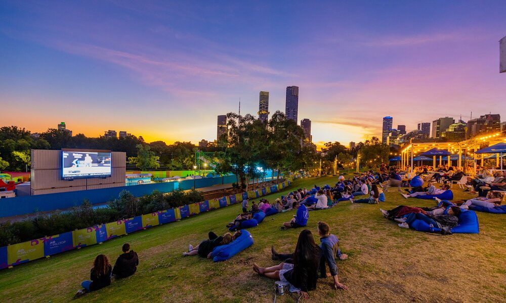 People sitting on blue beanbags on a grassy hill watching tennis on a large screen with the city and sunset behind.