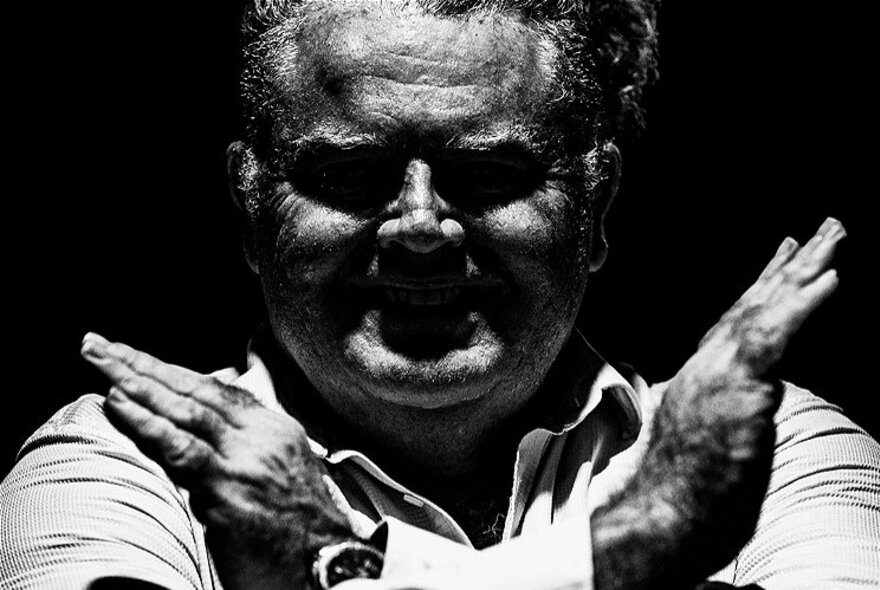 A black and white image of a man's upper body with his face not quite in the light, both hands up in the X position. 