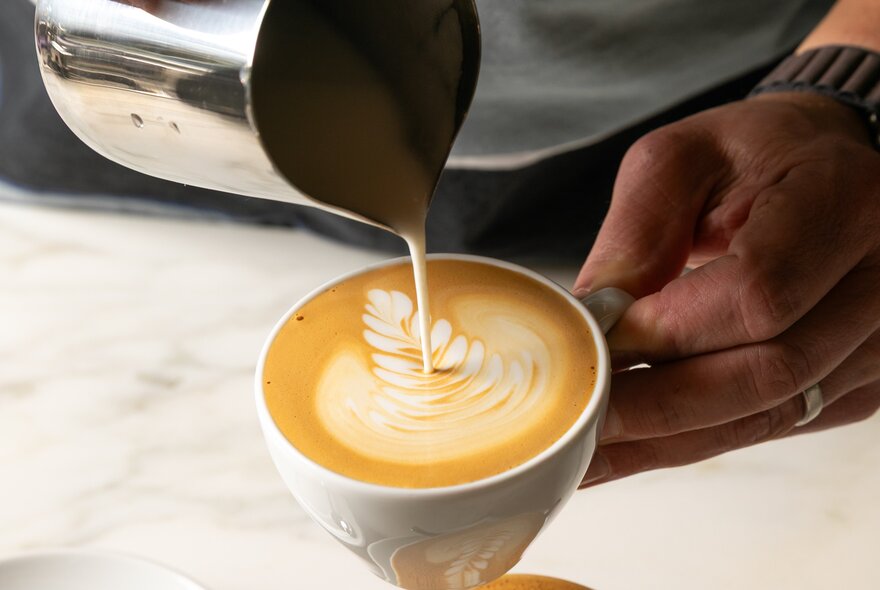 A person pouring warm milk from a jug into a cup of coffee and creating latte art.