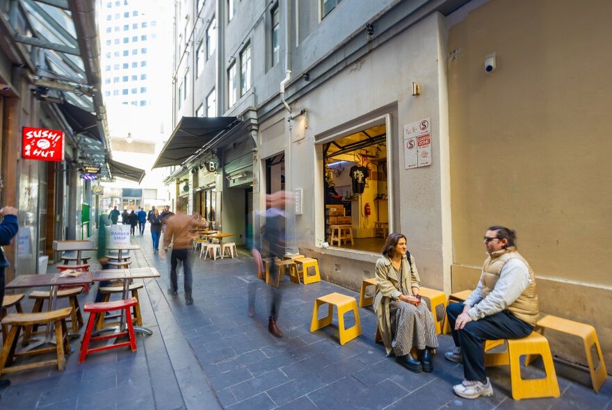 Two people sitting on yellow stools outside a busy laneway cafe.
