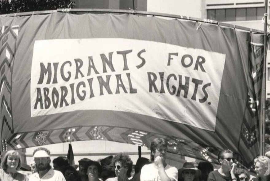 A protest banner from the 1960s with the slogan 'Migrants for Aboriginal Rights' and people marching underneath it.