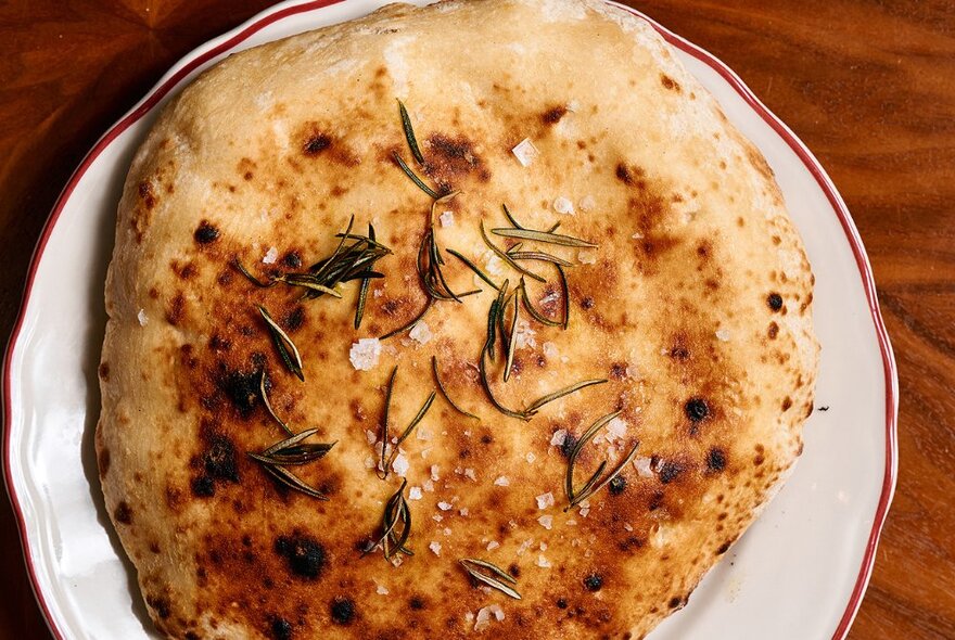 A woodfired flatbread on a white plate, with rosemary and salt flakes on it.