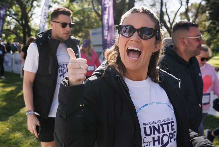 Woman wearing sunglasses and an event logo T-shirt giving the thumbs up, with other attendees in the background.