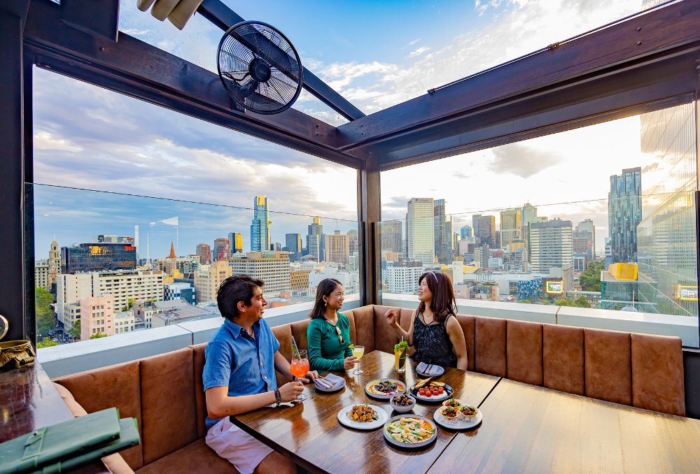 Three friends with share plates and cocktails in the corner booth of a city rooftop bar.