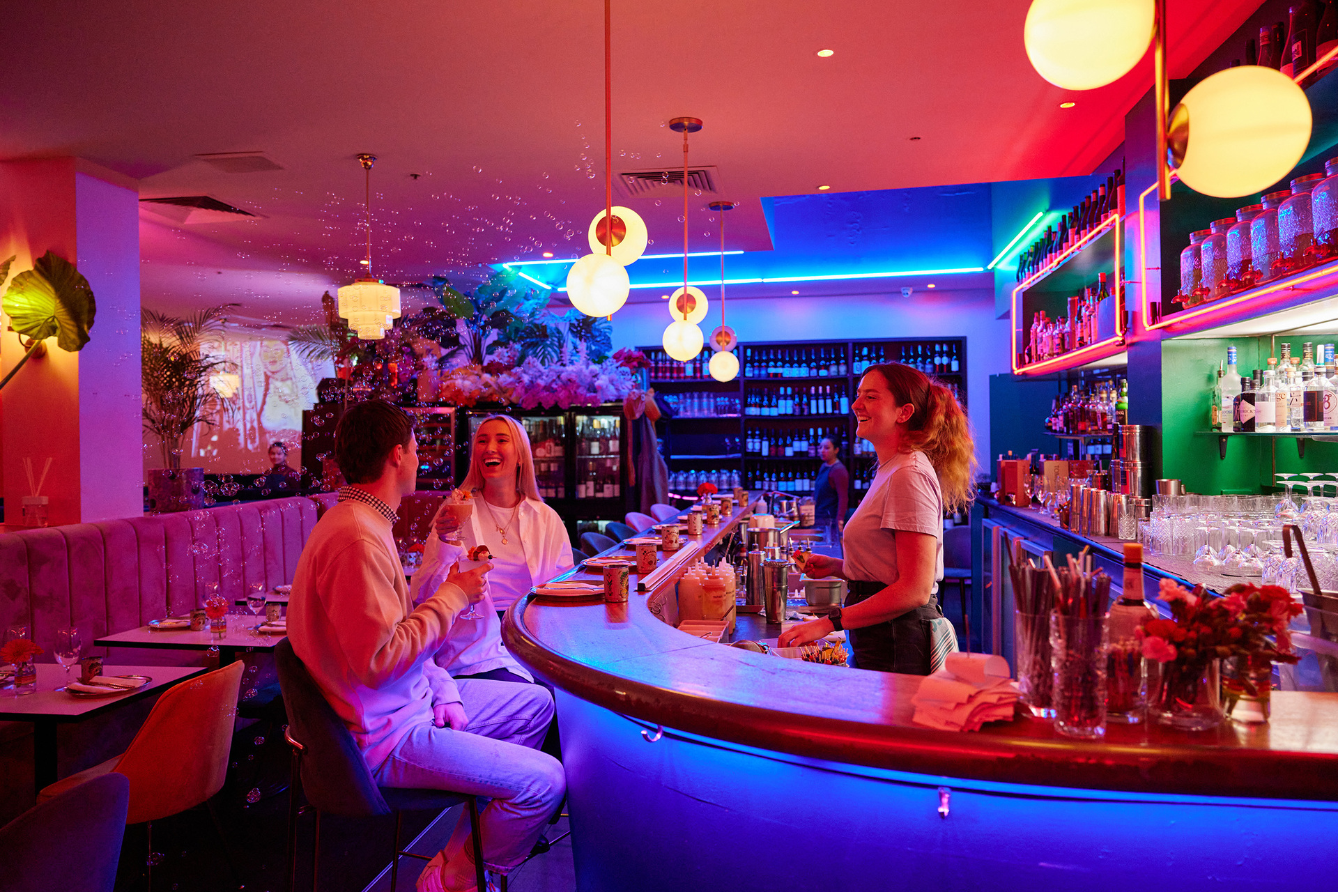 Two friends are sitting at a neon-lit bar talking with the bar tender