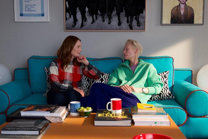 Two actors seated on a blue couch in conversation, coffee cups and books on a table in front of them.