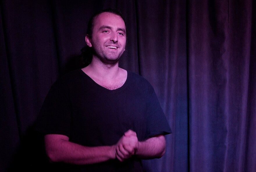 Comedian Joe Eidelson, grinning with his hands clasped together in front of a stage curtain. 