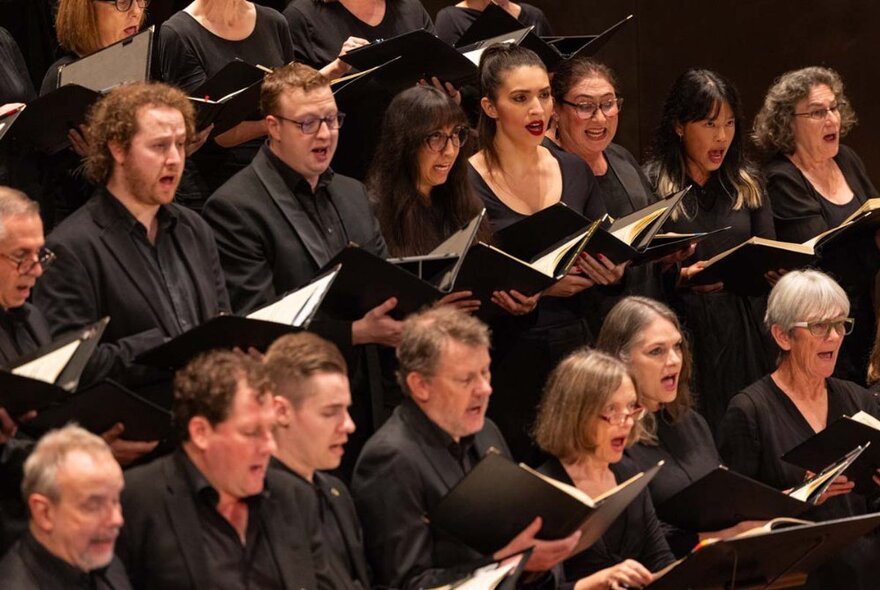 Massed choir standing in rows singing, wearing black shirts and holding open music books.