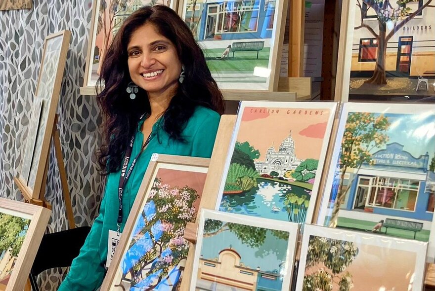 A smiling woman standing behind brightly coloured artworks of recognisable Melbourne scenes.