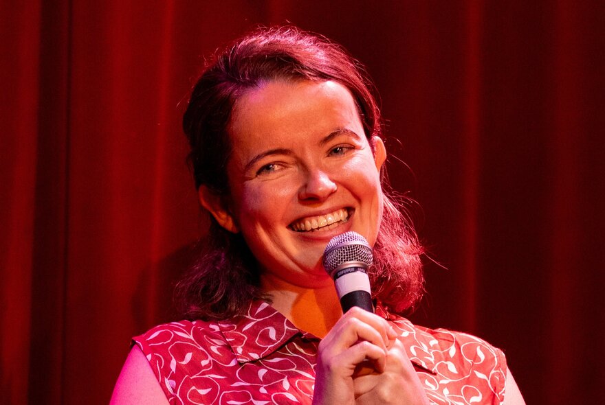 Comedian Roisin McAlinden smiling as she talks into a microphone on stage.