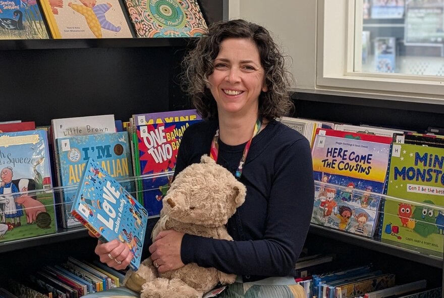 A woman holding a teddy about toe read a picture book in a library story area. 