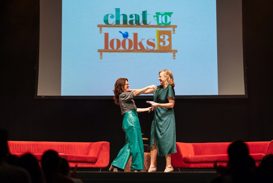 Annabel Crabb and Leigh Sales walking towards each other on a theatre stage to hug, behind them are two red couches and a screen with the words 'Chat 10 looks 3' displayed.