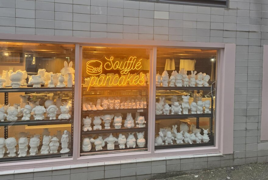 Exterior window of a cafe with shelves of small white plaster figures on display, a neon sign on the window reads 'Souffle Pancakes'.