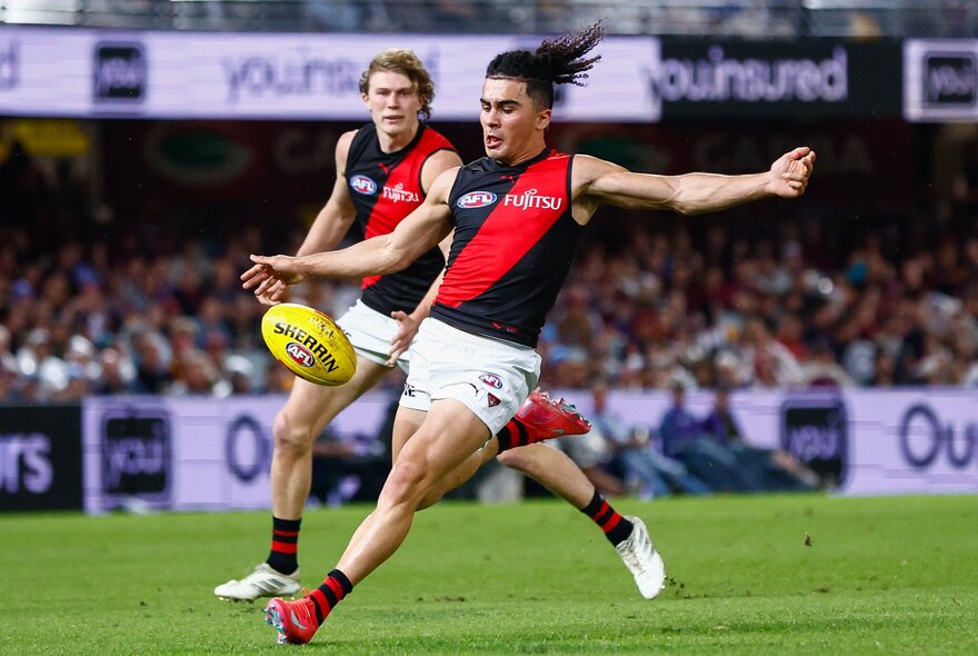 An Essendon AFL player running on the field about to kick a yellow football, with another Essendon player and the blurred stadium crowd behind him.