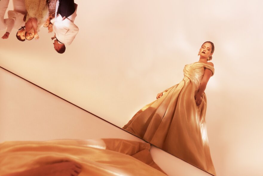 A woman in a ball gown standing by herself, her eyes downcast, in the top left corner of the image but upside down  is a group of people socialising.