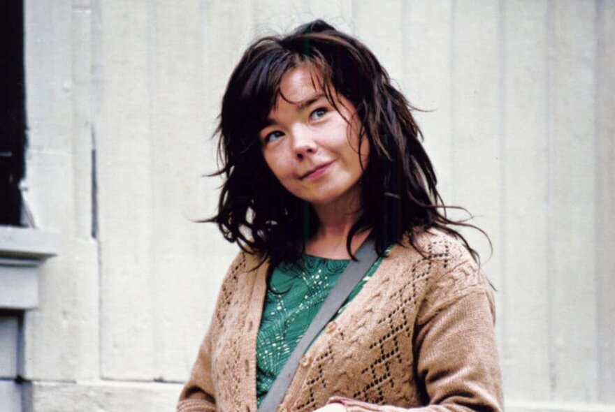 Singer and musician Björk smiling at the camera wearing a cardigan with her head tilted to one side. 