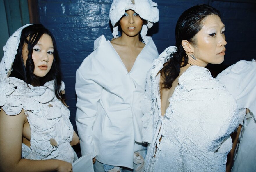 Three fashion models, all wearing white garments, getting ready to walk in a runway show.