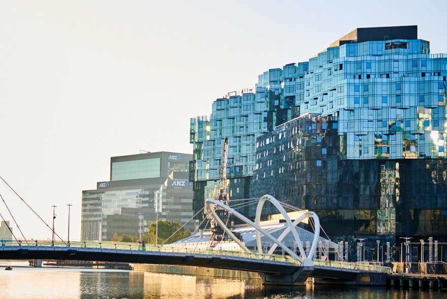 A large building with a glass exterior on the banks of the Yarra River, a pedestrian bridge in the foreground.