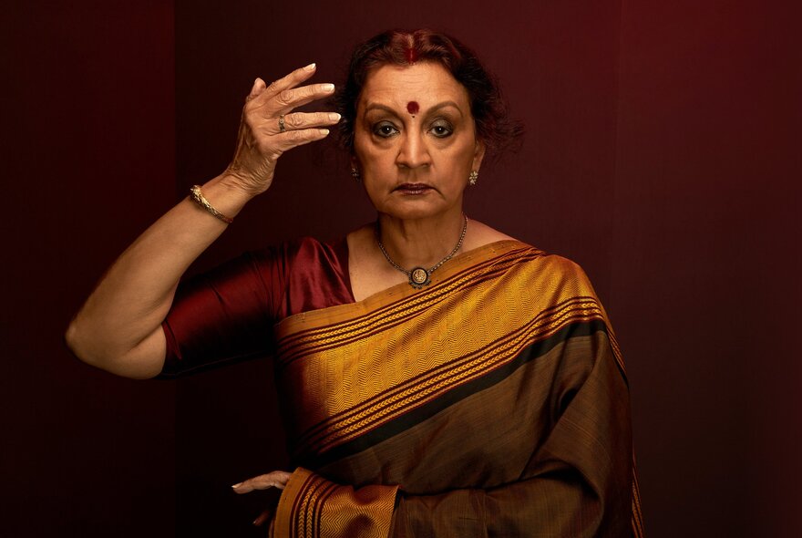 A woman with dark hair pulled back, wearing a gold and maroon sari and bindi, looks intently at the camera with her right hand raised near her head.