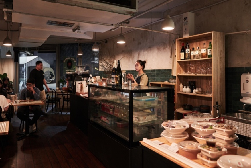 The interior of a small Japanese restaurant, showing a service area and staff, and an area with tables and chairs.