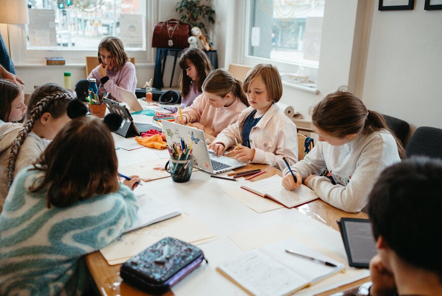 Kids seated around a large table writing and drawing during a creative writing workshop in a bright studio space.