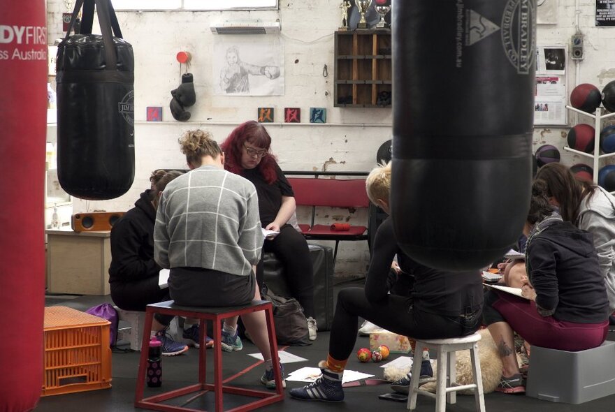 People sitting on chairs inside a boxing gym, writing in notebooks, surrounded by boxing equipment such as punching bags and boxing gloves.