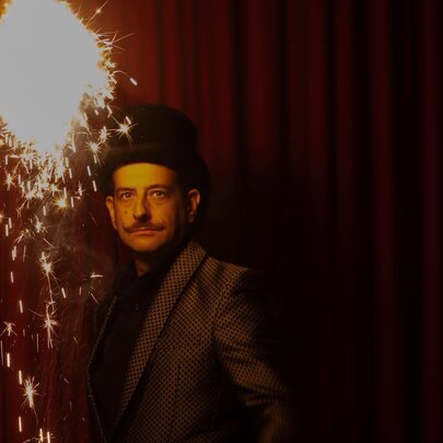 Magician and performer Anthony Demasi, wearing a black top hat and jacket, he stands under a oversize sparkler emitting light and sparkles.