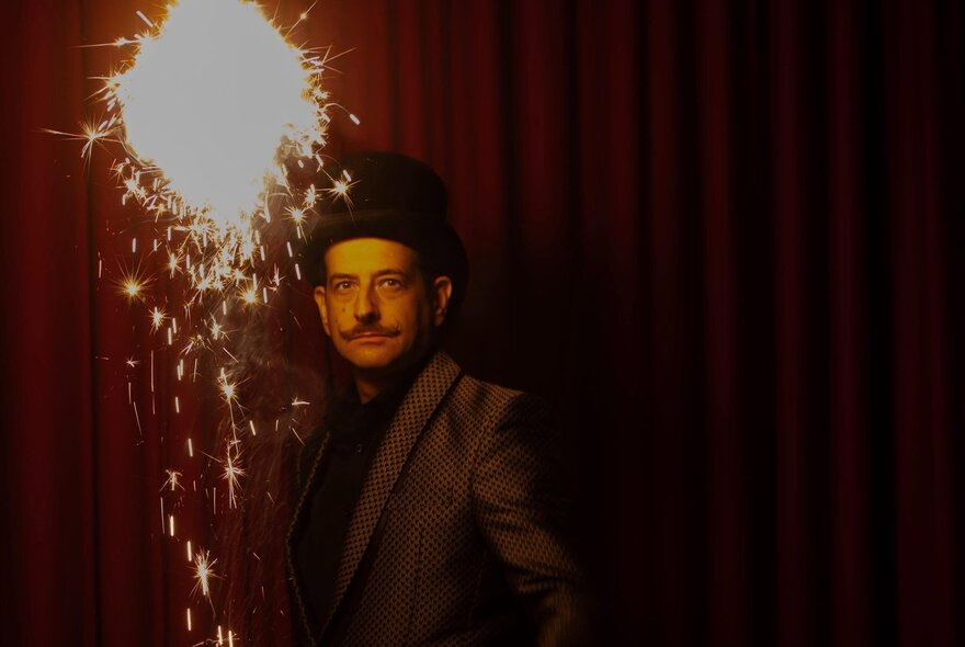Magician and performer Anthony Demasi, wearing a black top hat and jacket, he stands under a oversize sparkler emitting light and sparkles.