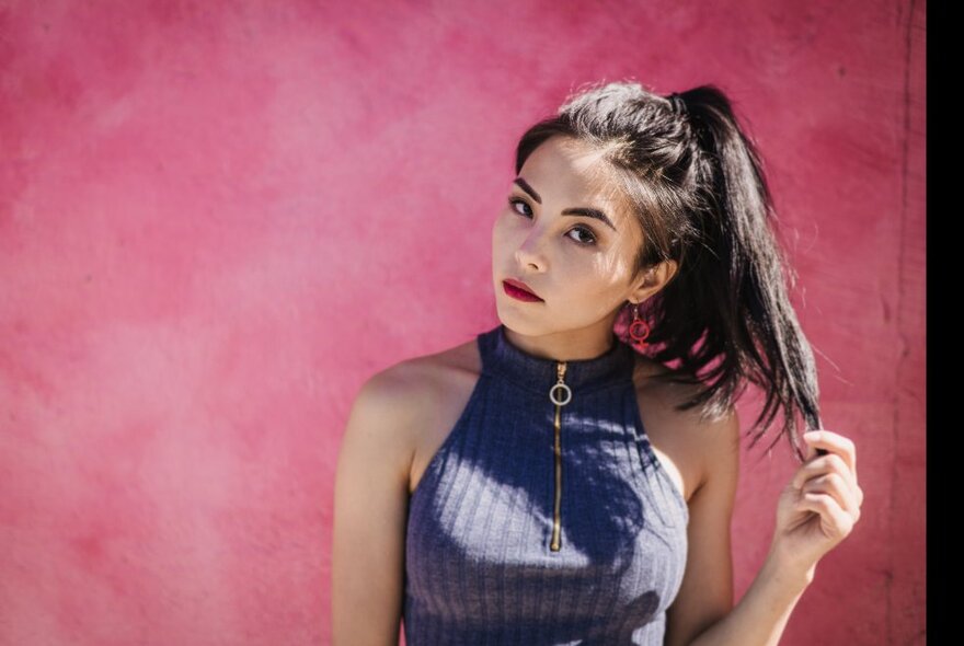 Woman holding the tip of her ponytail with her head on one side, against a pink wall.