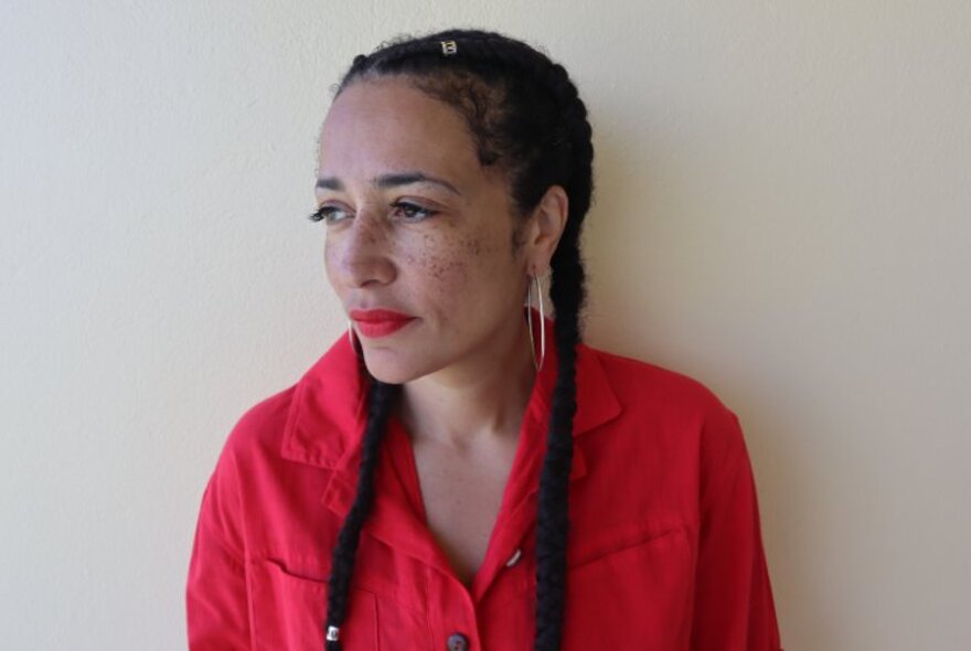 Portrait of acclaimed author, Zadie Smith, looking to her right and wearing a red shirt with her long black hair braided into two plaits.